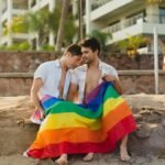 Two men sitting closely on a beach, wrapped in a vibrant rainbow flag, showcasing warmth and intimacy.