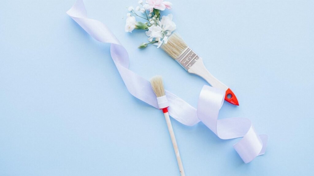 Two paint brushes with ribbon and flowers on a pastel blue background, ideal for DIY and decor themes.