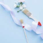 Two paint brushes with ribbon and flowers on a pastel blue background, ideal for DIY and decor themes.