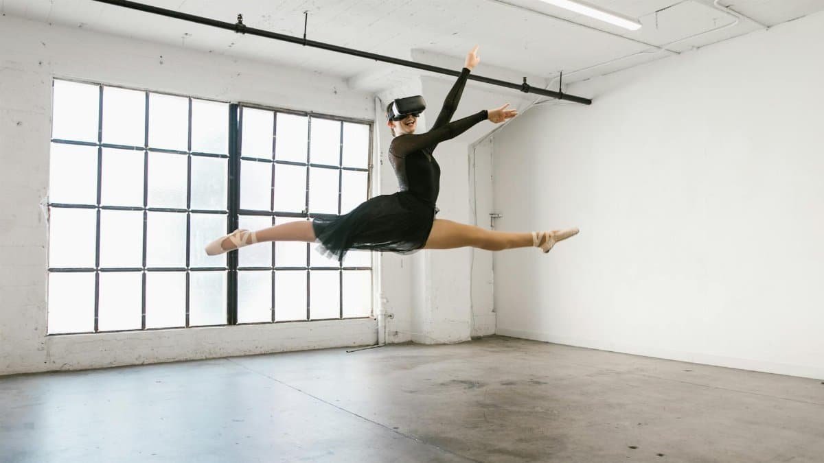 Ballet dancer wearing VR headset performs a leap in sunny studio, blending classical dance with modern technology.
