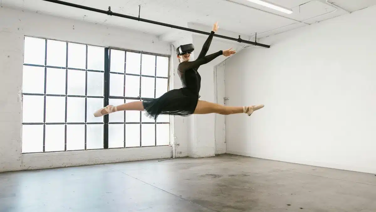 Ballet dancer wearing VR headset performs a leap in sunny studio, blending classical dance with modern technology.