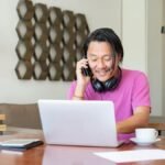 Man multitasking with phone and laptop in a cozy home setting, enjoying remote work flexibility.