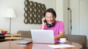 Man multitasking with phone and laptop in a cozy home setting, enjoying remote work flexibility.