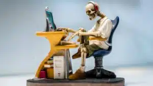 A quirky skeleton figurine wearing headphones sits at a desk with a computer, symbolizing burnout.