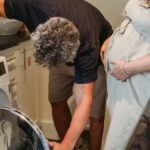 Crop pregnant female standing in kitchen while faceless male with gray curly hair helping to turn on washing machine and preparing laundry