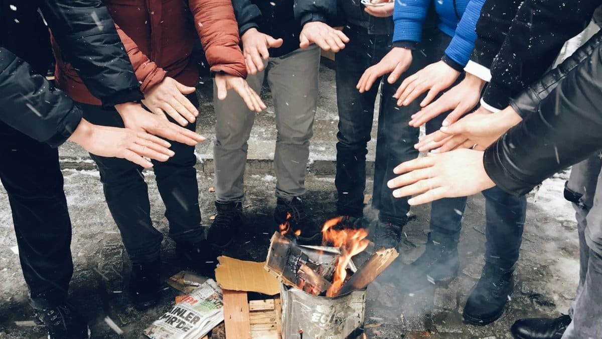 A group of people huddled together warming their hands over an outdoor fire during winter.