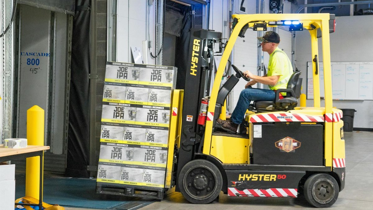 A worker drives a Hyster forklift moving Joe IPA boxes in a warehouse.