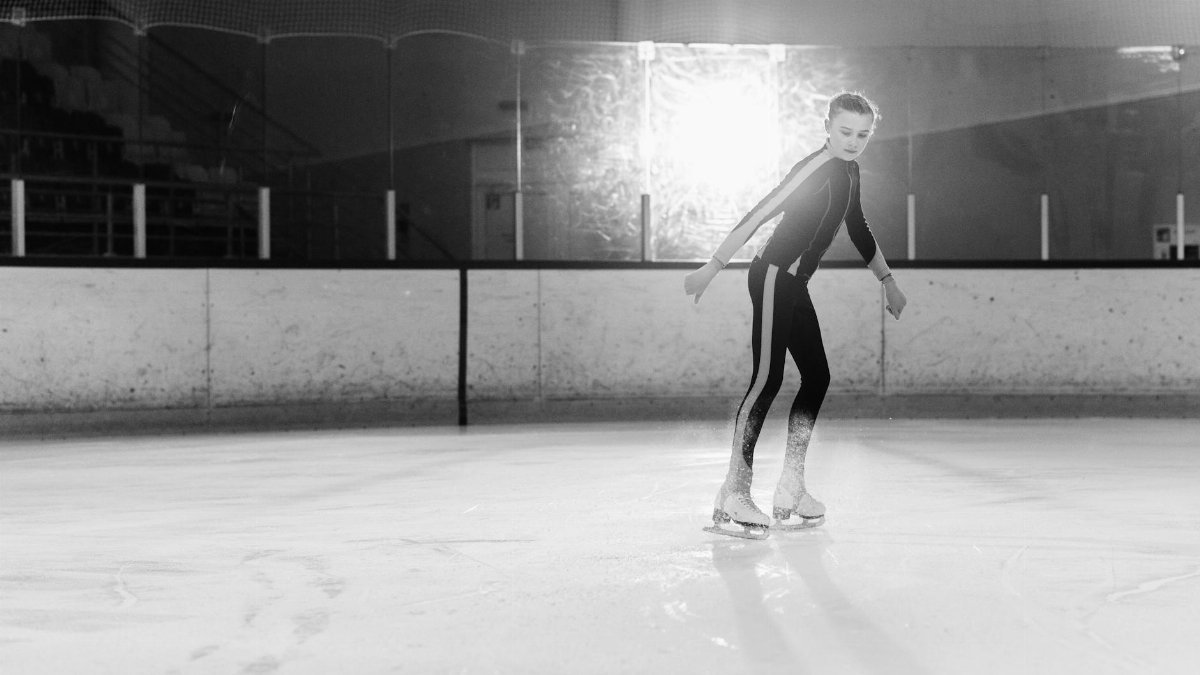 Black and white image of a girl figure skating on an ice rink indoors.