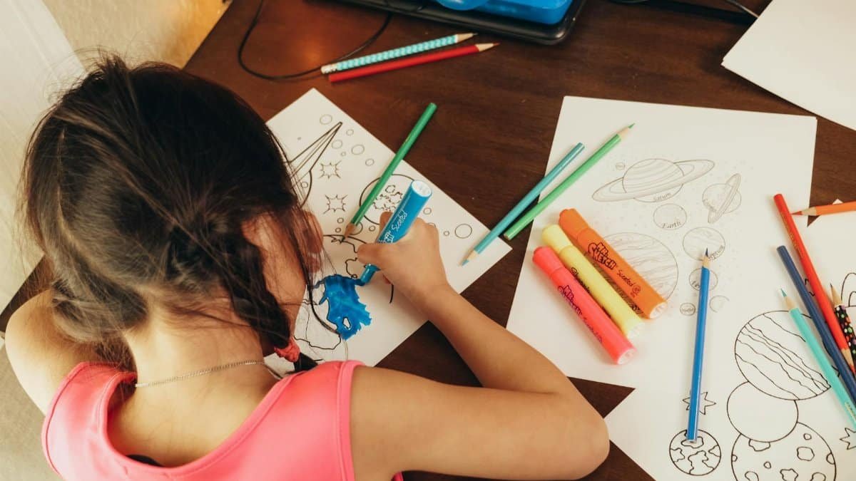 Young girl coloring space-themed drawings with markers and pencils indoors.