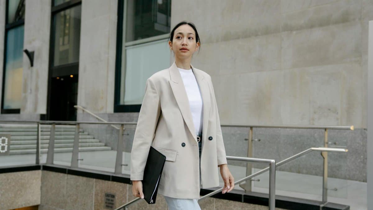 Confident Asian female entrepreneur in stylish outfit and with folder walking in city street and looking away
