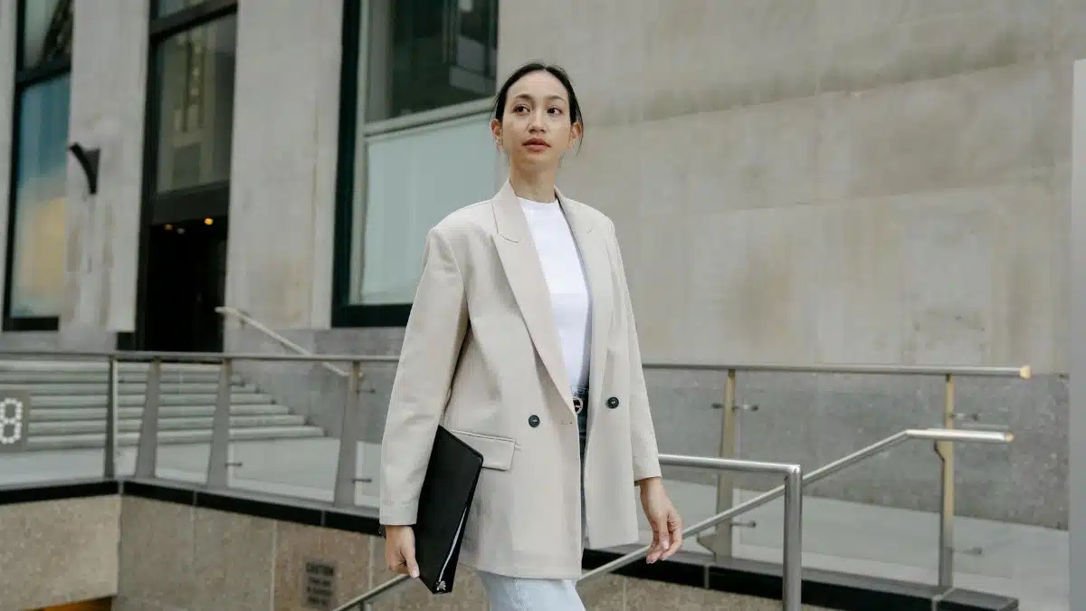 Confident Asian female entrepreneur in stylish outfit and with folder walking in city street and looking away