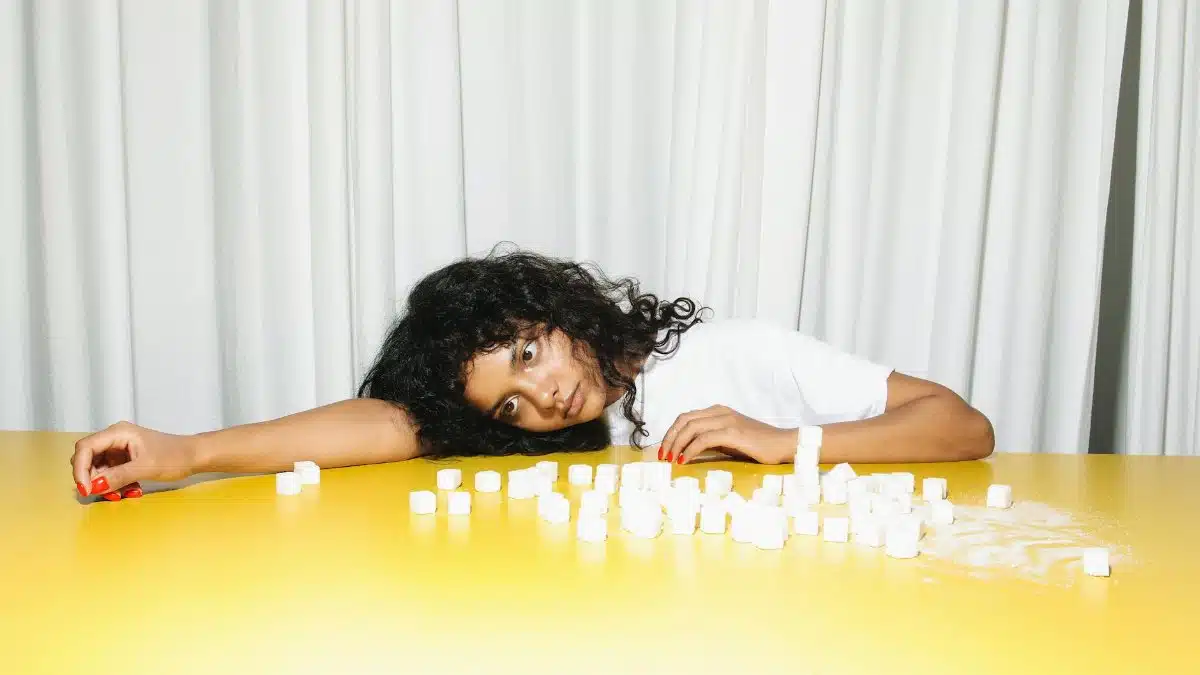 A woman leaning on a yellow table surrounded by sugar cubes, reflecting on health or lifestyle choices.