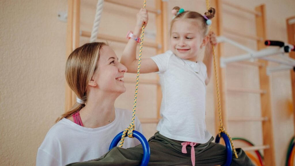 Smiling mother teaches her daughter gymnastics on rings indoors, fostering bonding and flexibility.