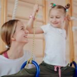 Smiling mother teaches her daughter gymnastics on rings indoors, fostering bonding and flexibility.