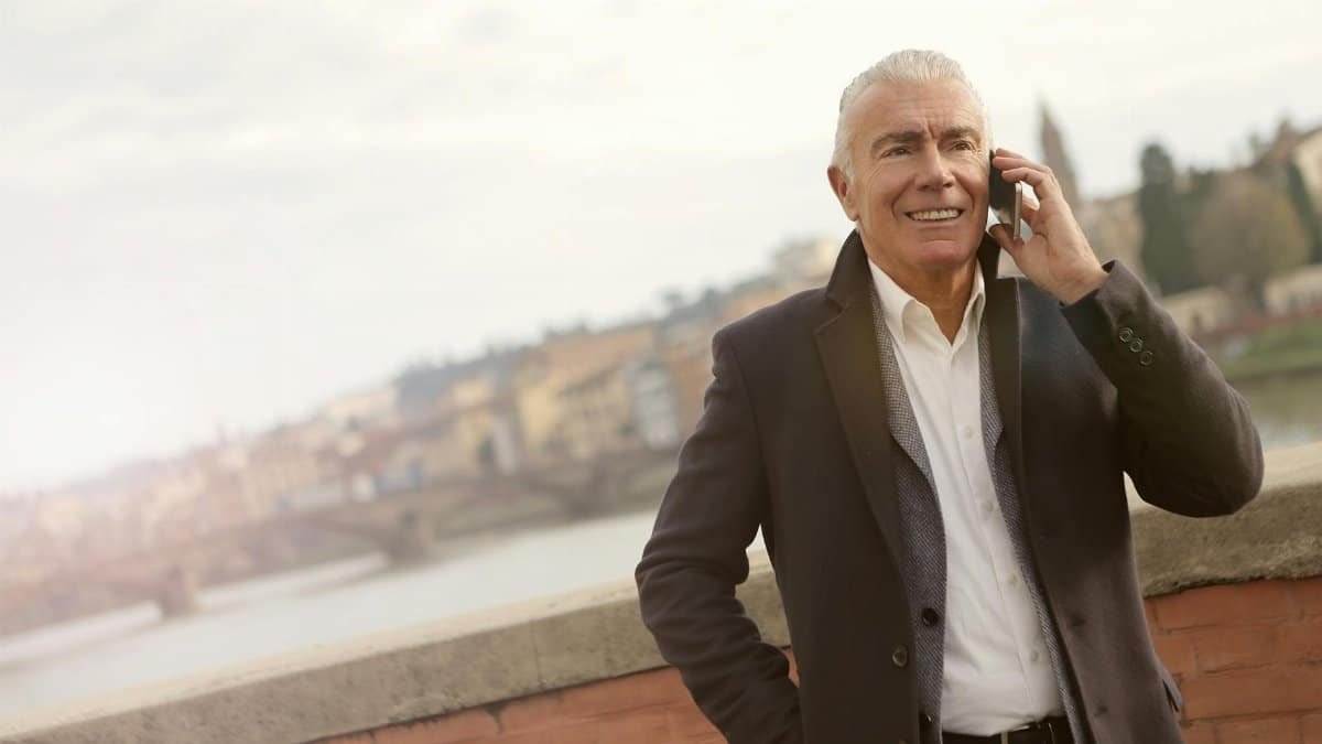 Senior man wearing business coat talking on mobile phone with scenic city backdrop.