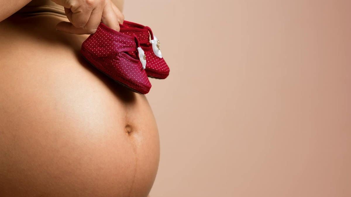 Close-up of a pregnant woman holding red baby shoes on her belly, symbolizing anticipation and love.
