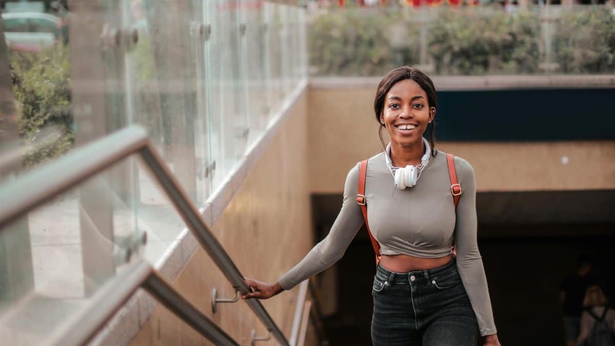 A cheerful young woman with headphones walks confidently outdoors down the steps.