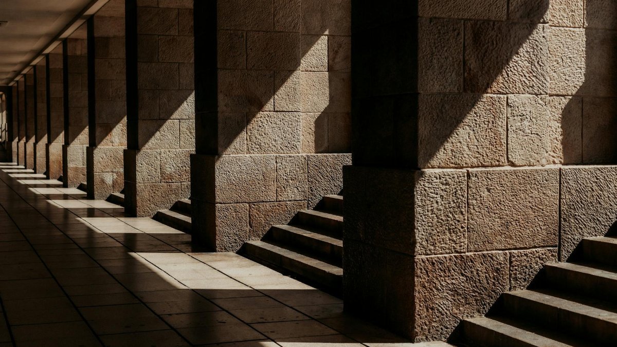 Sunlight creates patterns on stone columns and steps in this architectural corridor scene.