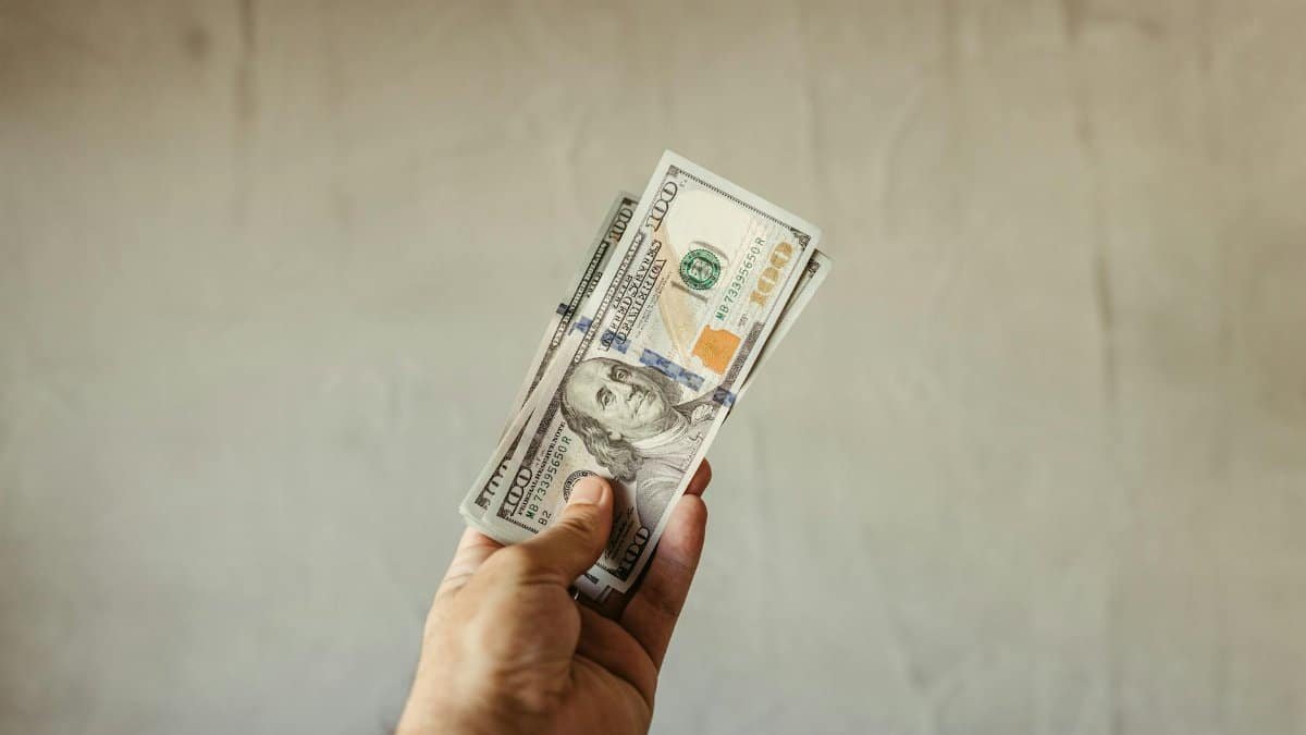 A hand holding a stack of US dollar banknotes against a neutral backdrop, symbolizing wealth.