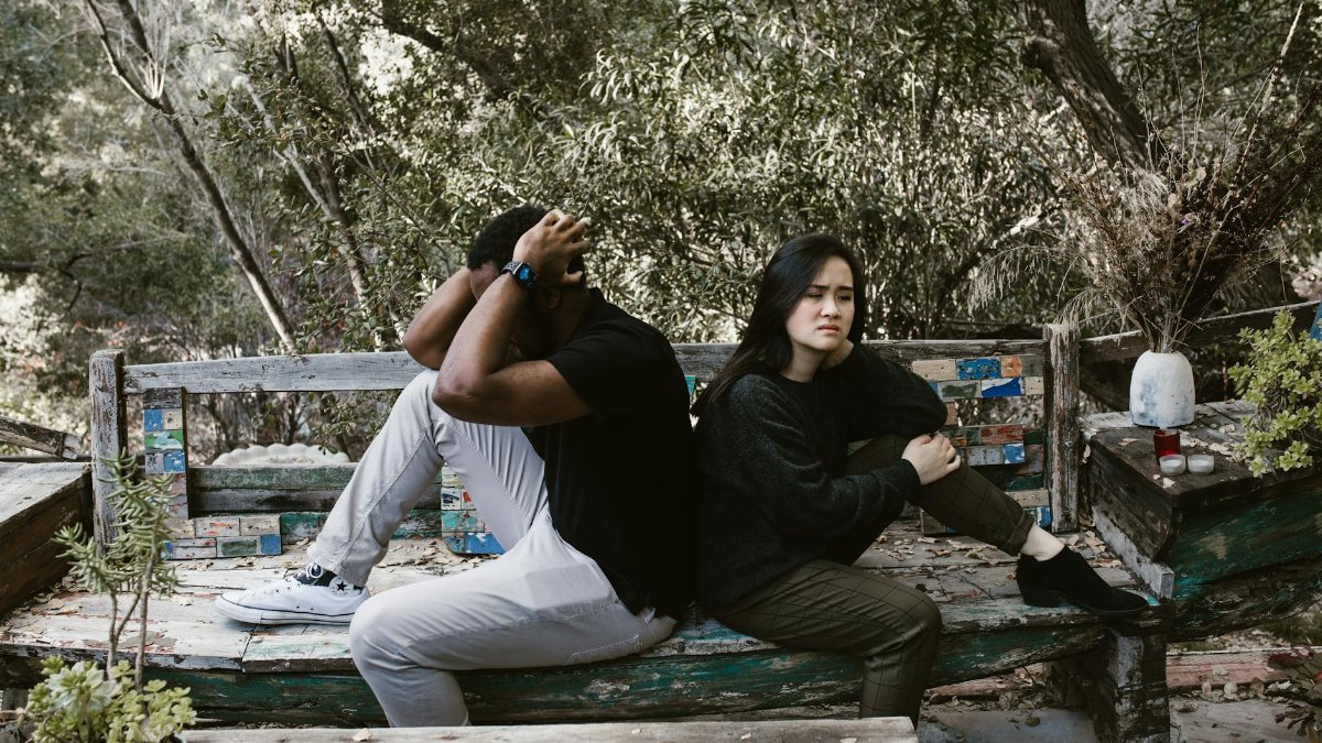 A diverse couple sits on a wooden bench outdoors, expressing anger and sadness.