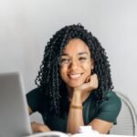 Joyful businesswoman with curly hair smiling at camera while using laptop indoors.