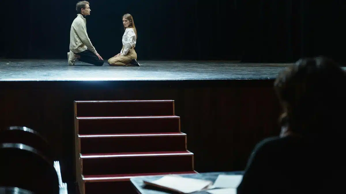 Actors in a dramatic rehearsal on stage, under dim theater lights.