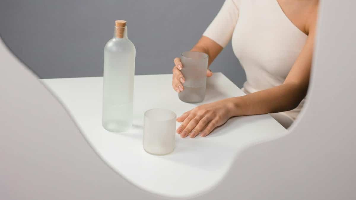 Elegant minimalist composition featuring a frosted glass bottle and drinking glass with a woman's hand.
