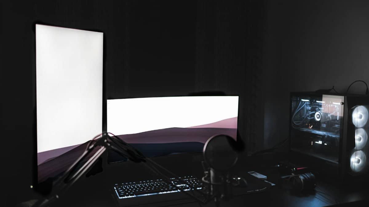 Gaming setup with glowing modern monitors placed on black table with keyboard near microphone and system unit in dark room