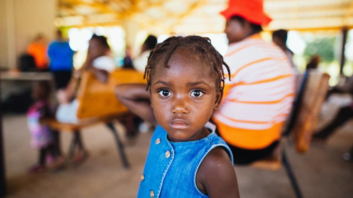 Cute African American girl with brown eyes and hand behind head looking at camera while standing in cafe