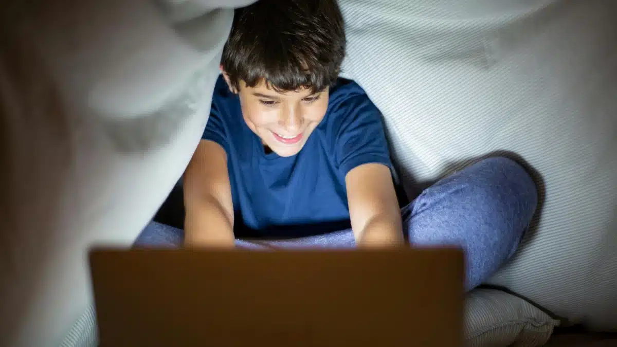 Smiling boy sitting under blanket using laptop, indoor fun at night.