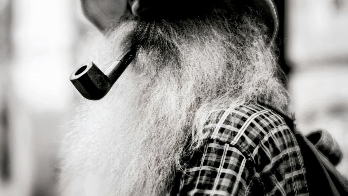 Close-up black and white portrait of an elderly man with a long beard, wearing a plaid shirt and hat, smoking a pipe.