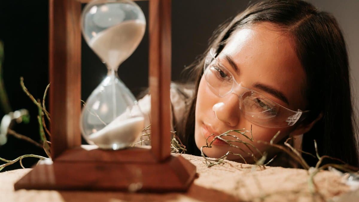 A woman wearing glasses observes an hourglass, symbolizing time and reflection.