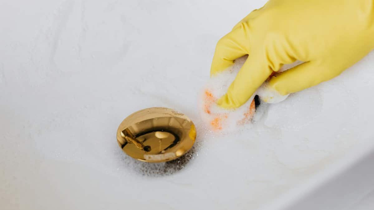 Close-up of a hand in a glove cleaning a foam-covered gold drain in a sink.