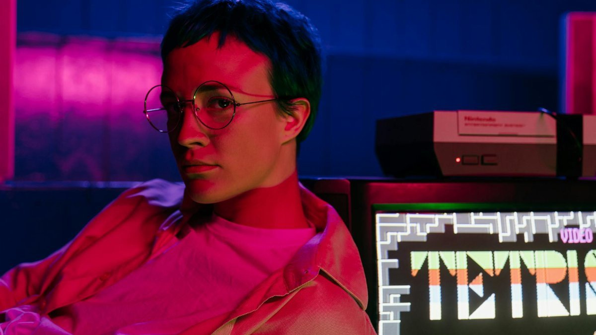 A teenager with eyeglasses in an 80s-style arcade displaying a Tetris game on screen.
