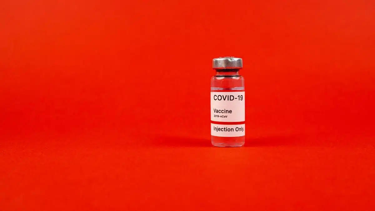 Glass vial of COVID-19 vaccine on a vibrant red backdrop, emphasizing protection.