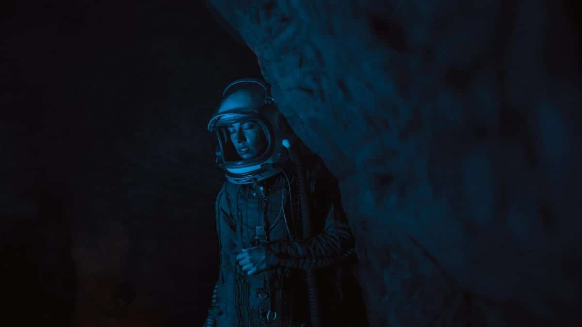 A person in an astronaut suit stands within a shadowy rock formation, atmospheric and contemplative.