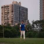 A teenage boy walks barefoot in a grassy urban park surrounded by tall buildings.