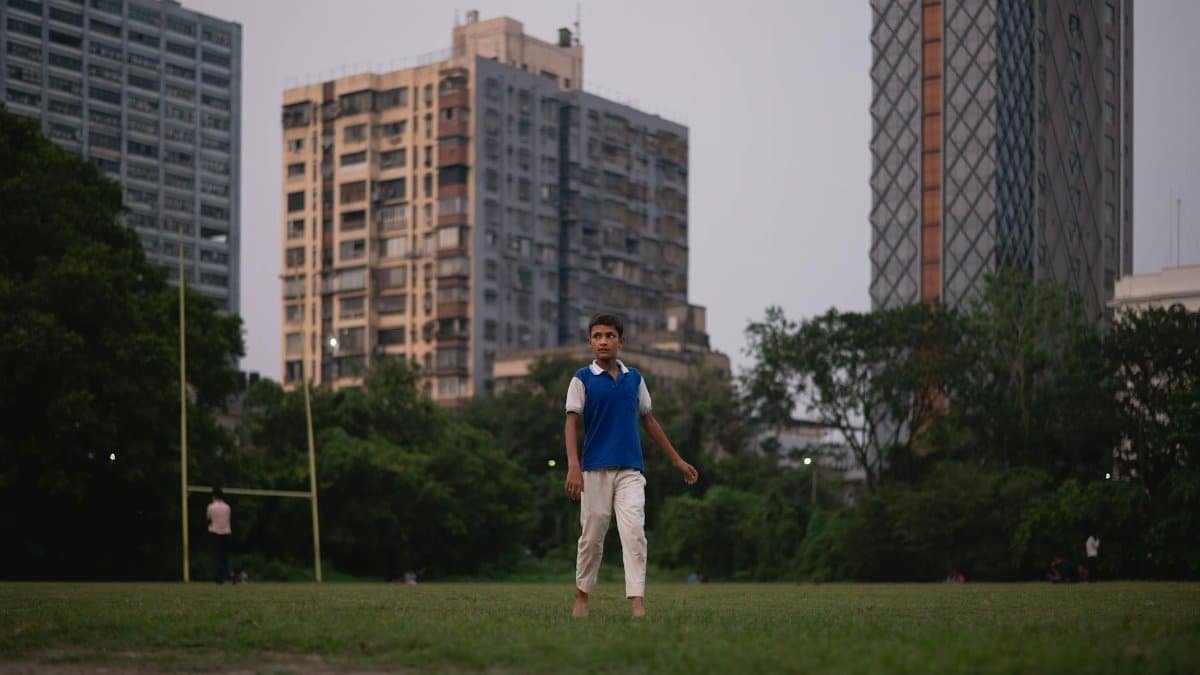 A teenage boy walks barefoot in a grassy urban park surrounded by tall buildings.