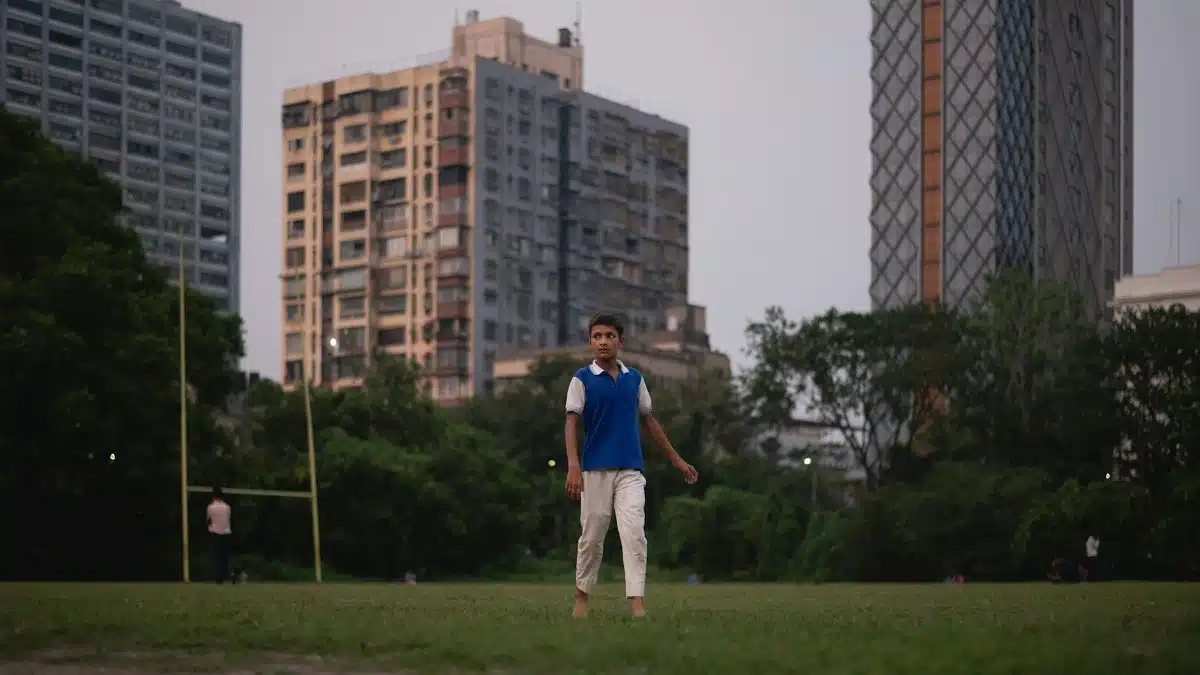 A teenage boy walks barefoot in a grassy urban park surrounded by tall buildings.