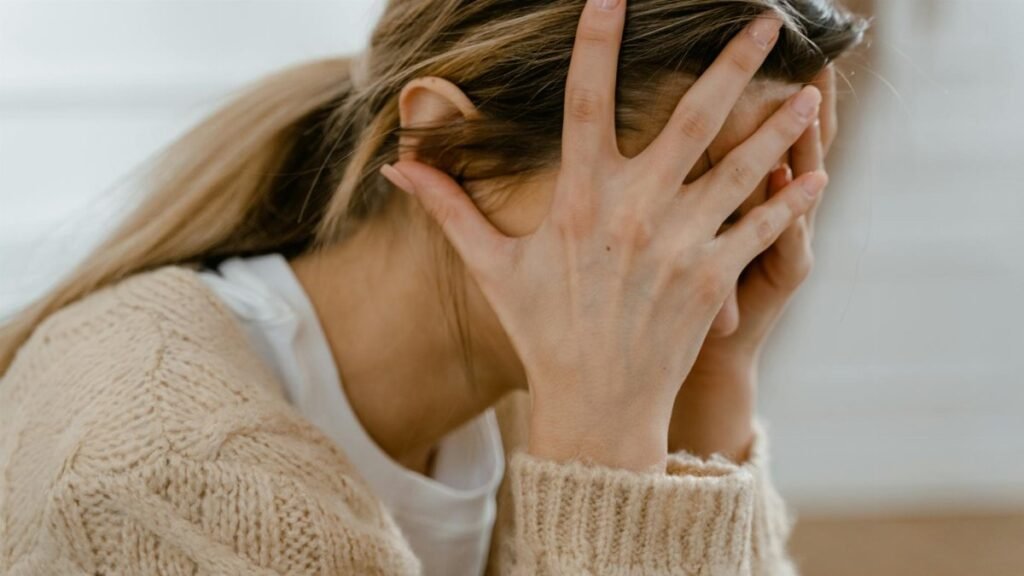 Woman sitting indoors with face covered by hands, expressing stress and frustration.