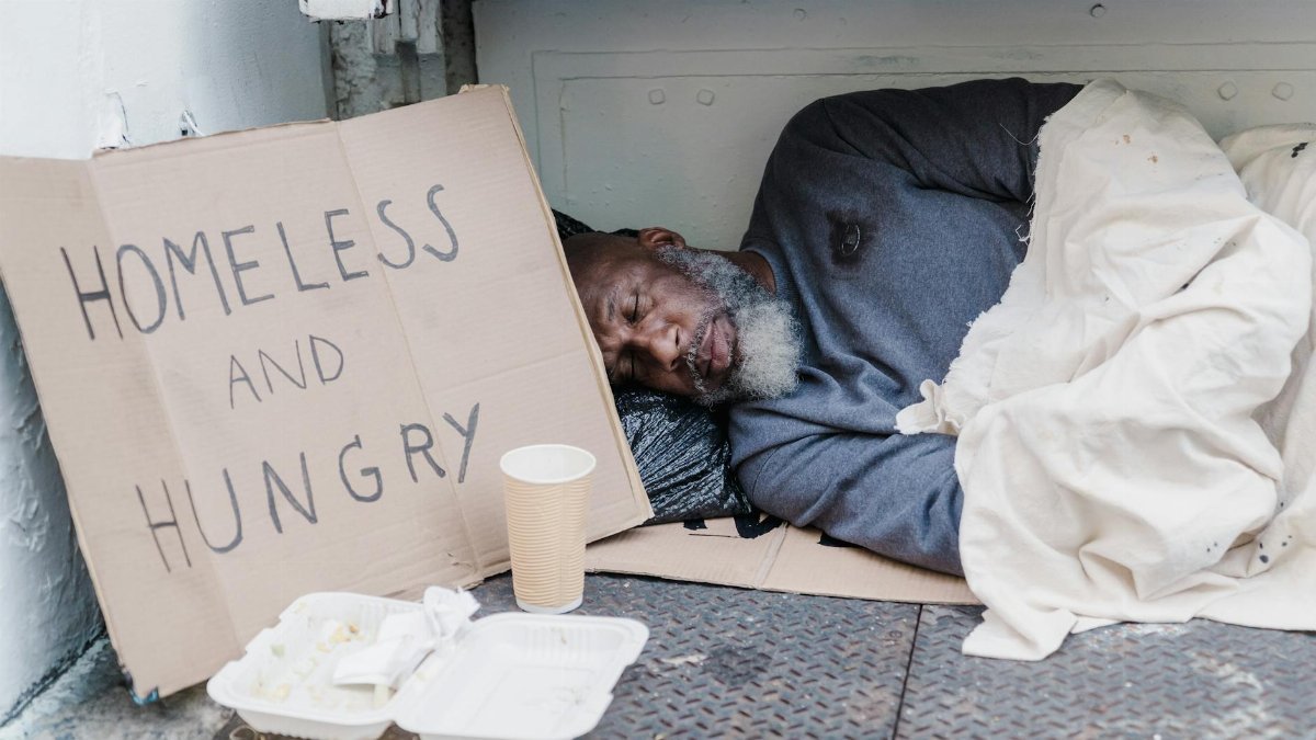 Elderly homeless man sleeping on the street with a sign reading 'Homeless and Hungry'.