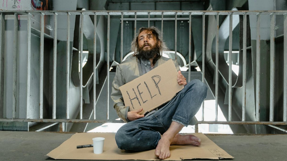 A homeless man sits under a bridge with a sign reading 'HELP', highlighting poverty and homelessness.