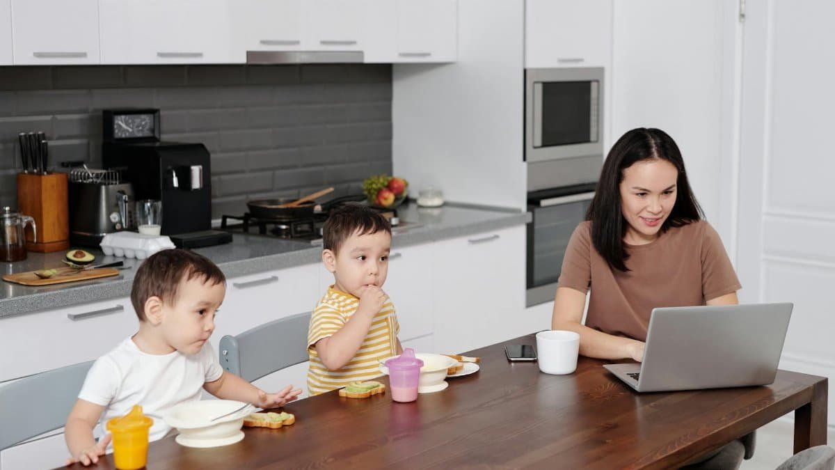 A mother multitasks on her laptop while her twin toddlers enjoy a snack in a modern kitchen setting.