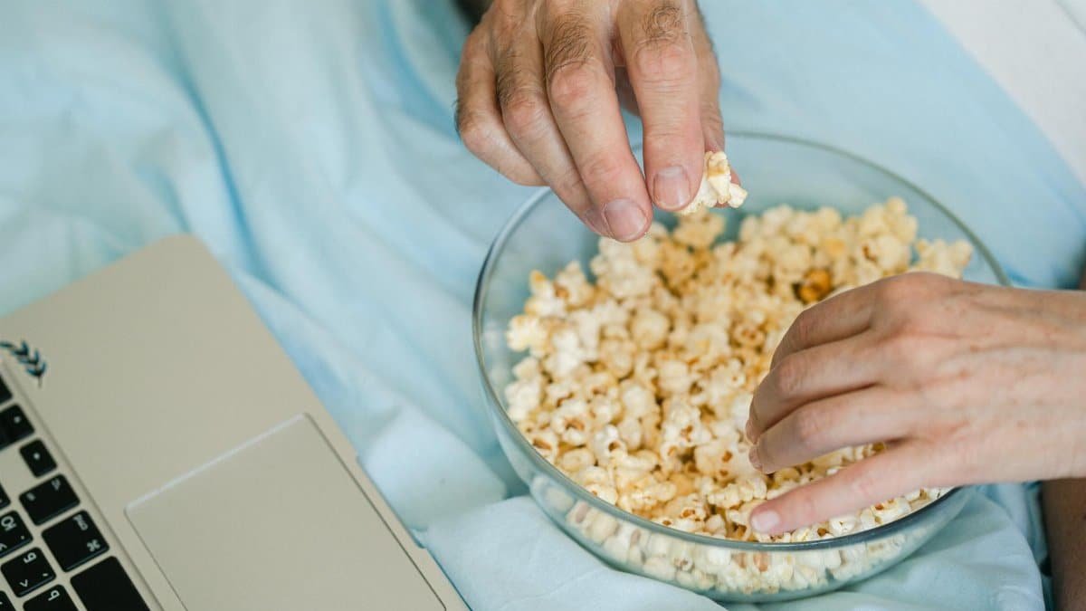 Hands reach for popcorn in a glass bowl next to a laptop, set for a cozy movie night at home.