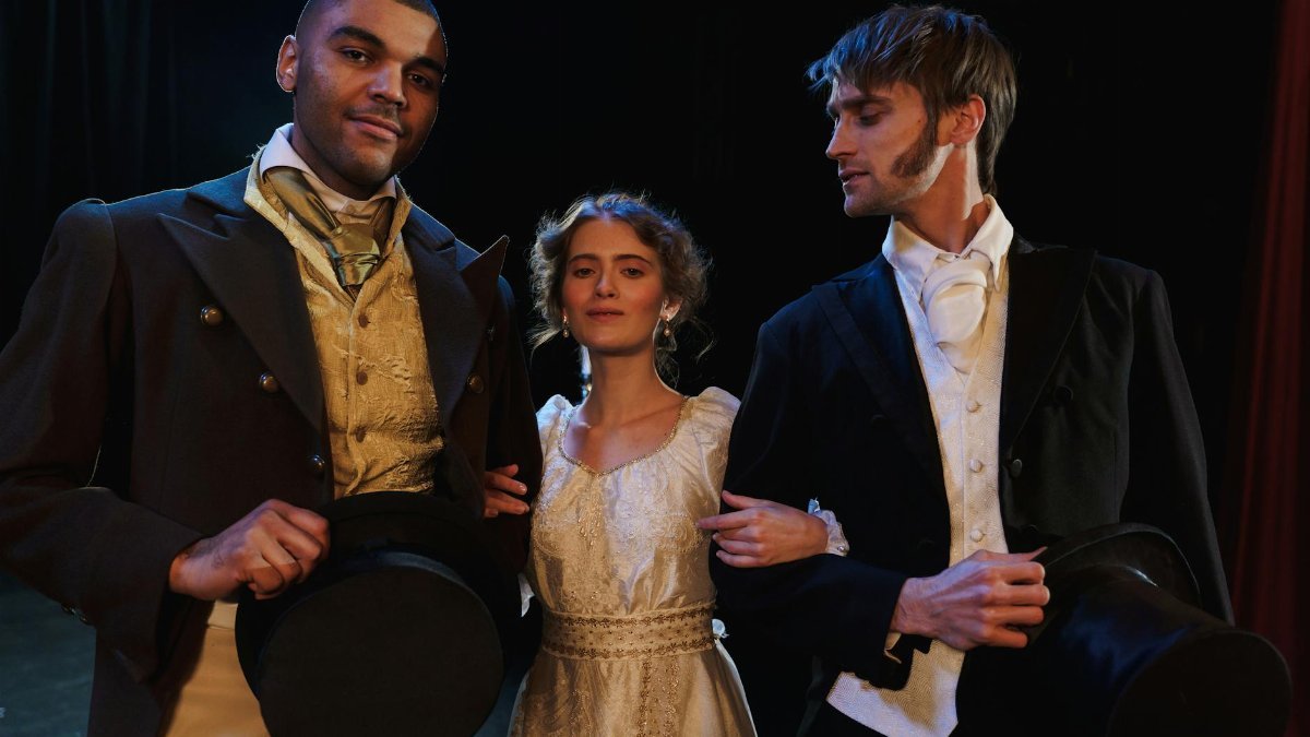 Actors in period costumes pose on stage, embodying classic theatrical drama.