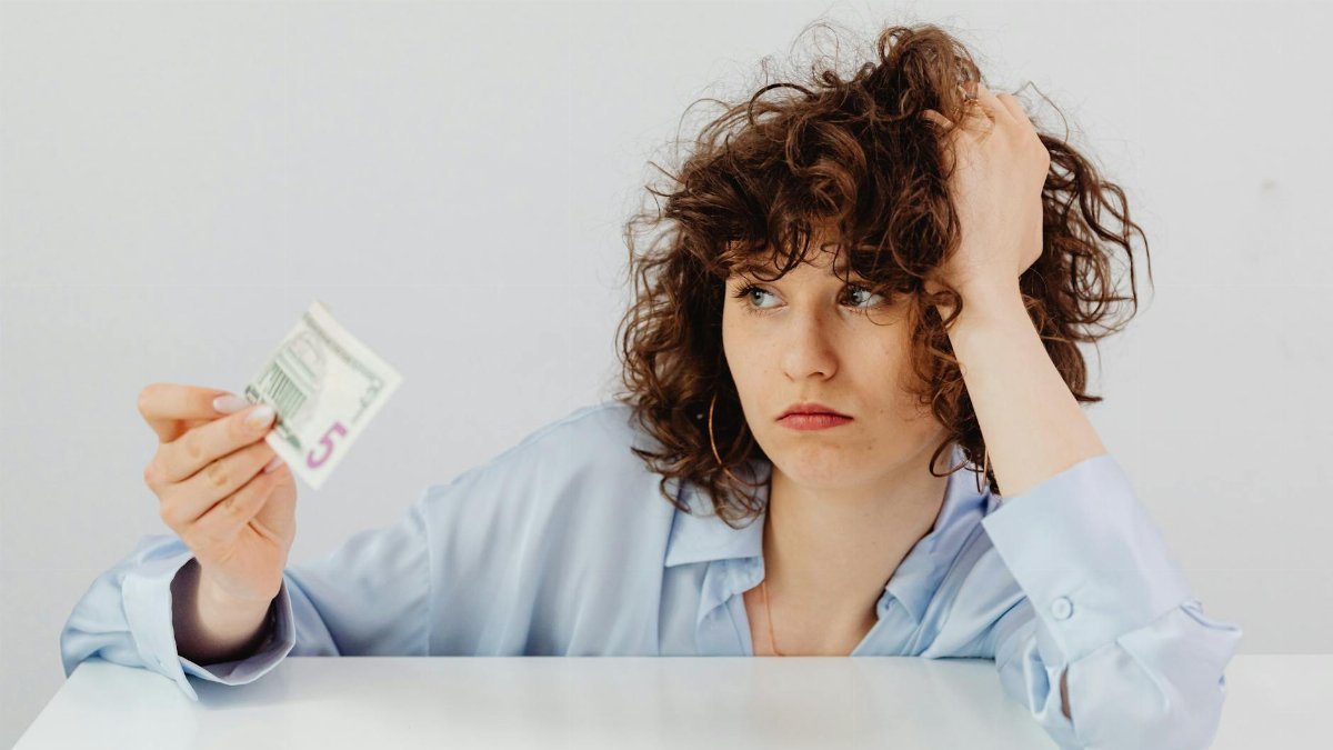 Concerned woman with curly hair holding a five dollar bill, expressing financial worry.