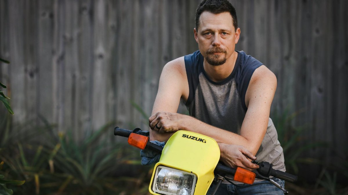Caucasian man posing with a yellow Suzuki motorcycle outdoors, showcasing a casual lifestyle vibe.