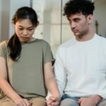 A couple sitting on a bed holding hands, facing relationship challenges indoors.