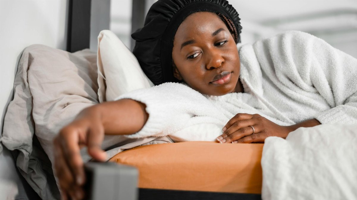 Woman in a cozy robe reaching for alarm clock in bedroom, waking up refreshed.