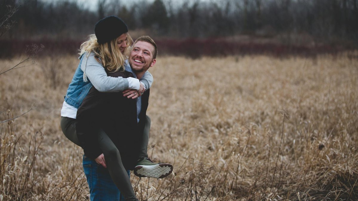 A happy couple enjoys a playful moment in a field during the day, symbolizing love and joy.